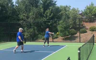 This is an image of two older white people playing pickleball wearing blue shirts in an outdoors sunny environment.